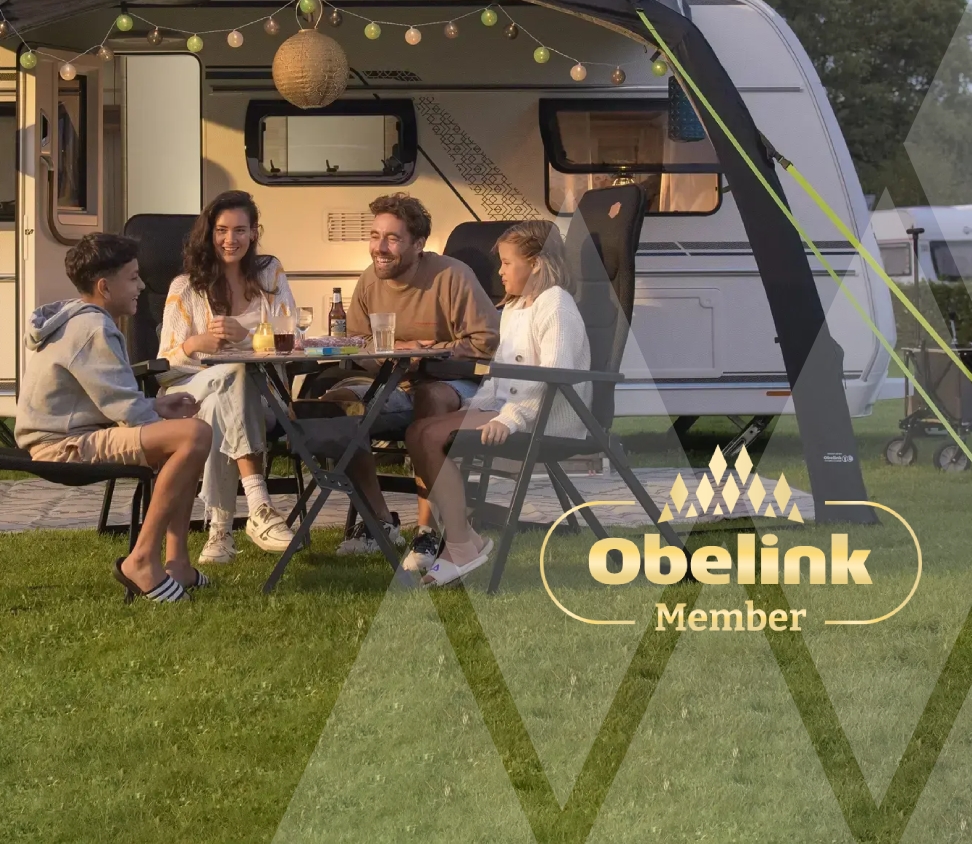 Wohnwagen mit Vorzelt, dekoriert mit stimmungsvoller Beleuchtung, Menschen genießen gemeinsam an einem Campingtisch in der Abenddämmerung auf einer Rasenfläche
