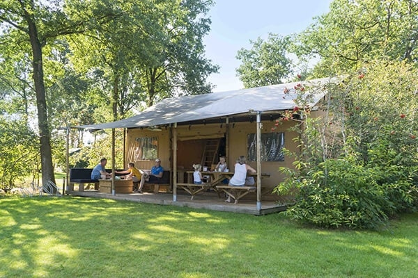 Luxuriöses Safarizelt mit überdachter Terrasse in grüner Umgebung, Picknicktisch aus Holz und Sitzgelegenheiten für Gäste an sonnigen Tagen