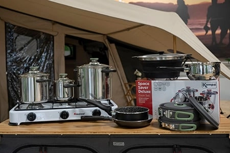 Campingkocher mit Töpfen und Pfannen auf Holztisch vor Zelt bei Sonnenuntergang, daneben verpacktes Kochgeschirr