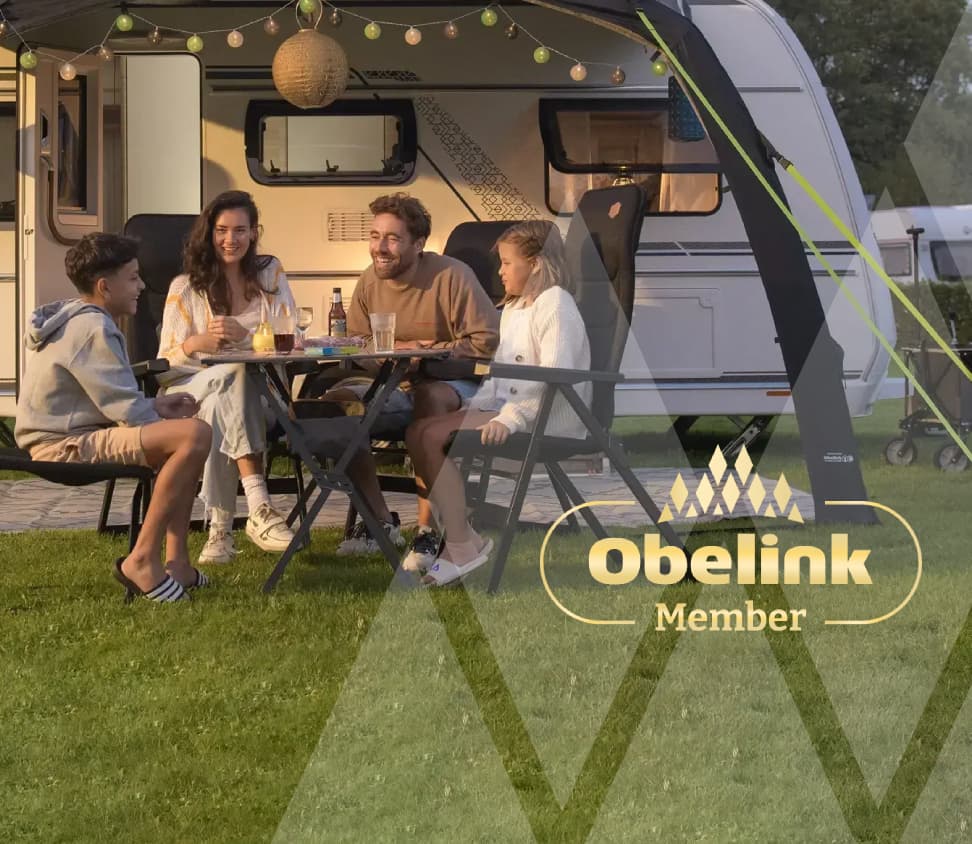 Wohnwagen mit Vorzelt, dekoriert mit stimmungsvoller Beleuchtung, Menschen genießen gemeinsam an einem Campingtisch in der Abenddämmerung auf einer Rasenfläche