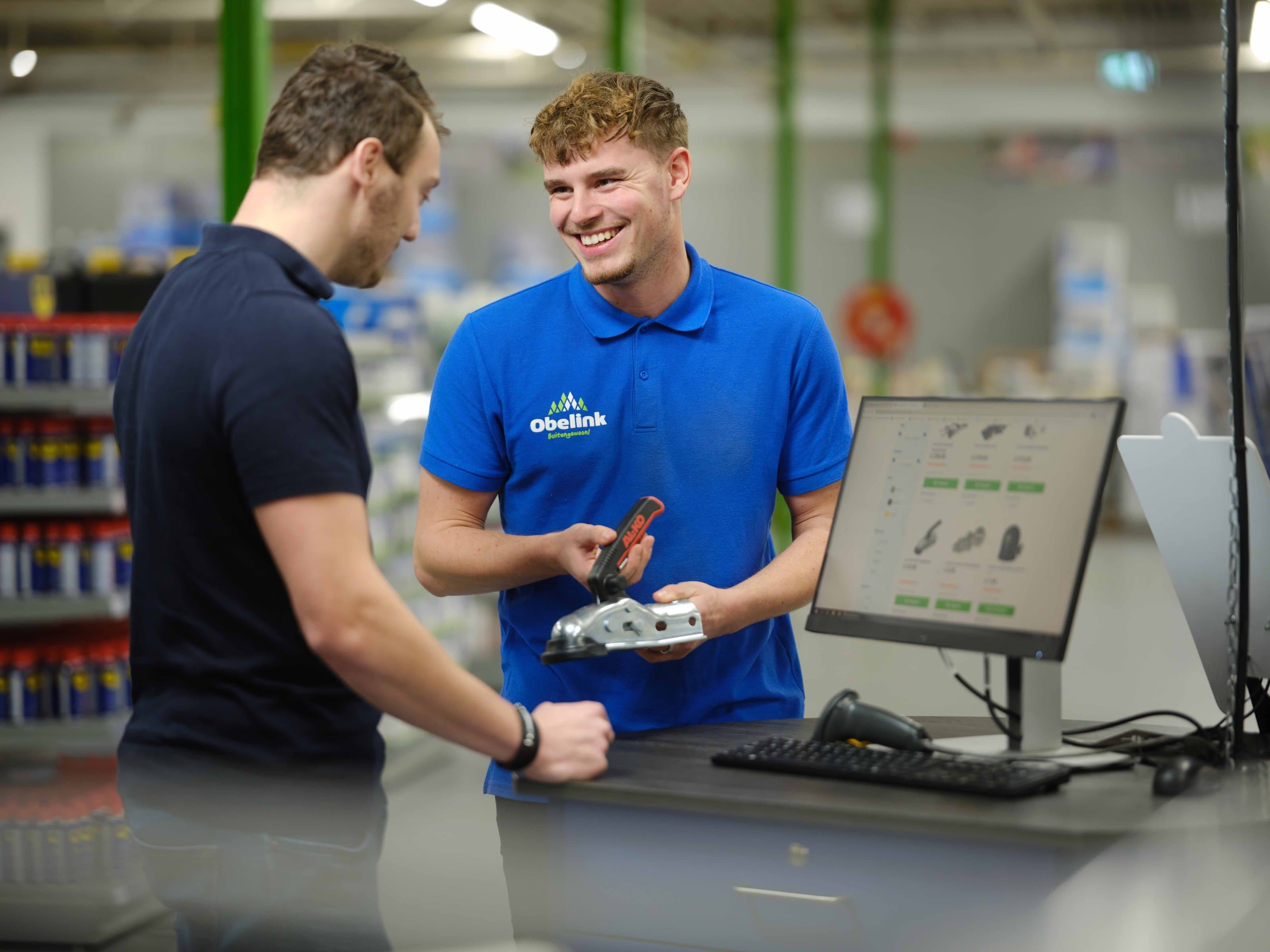 Mitarbeiter in blauem Obelink Poloshirt zeigt einem Kunden ein metallisches Befestigungsteil an der Verkaufstheke mit Computerbildschirm, auf dem Produkte sichtbar sind