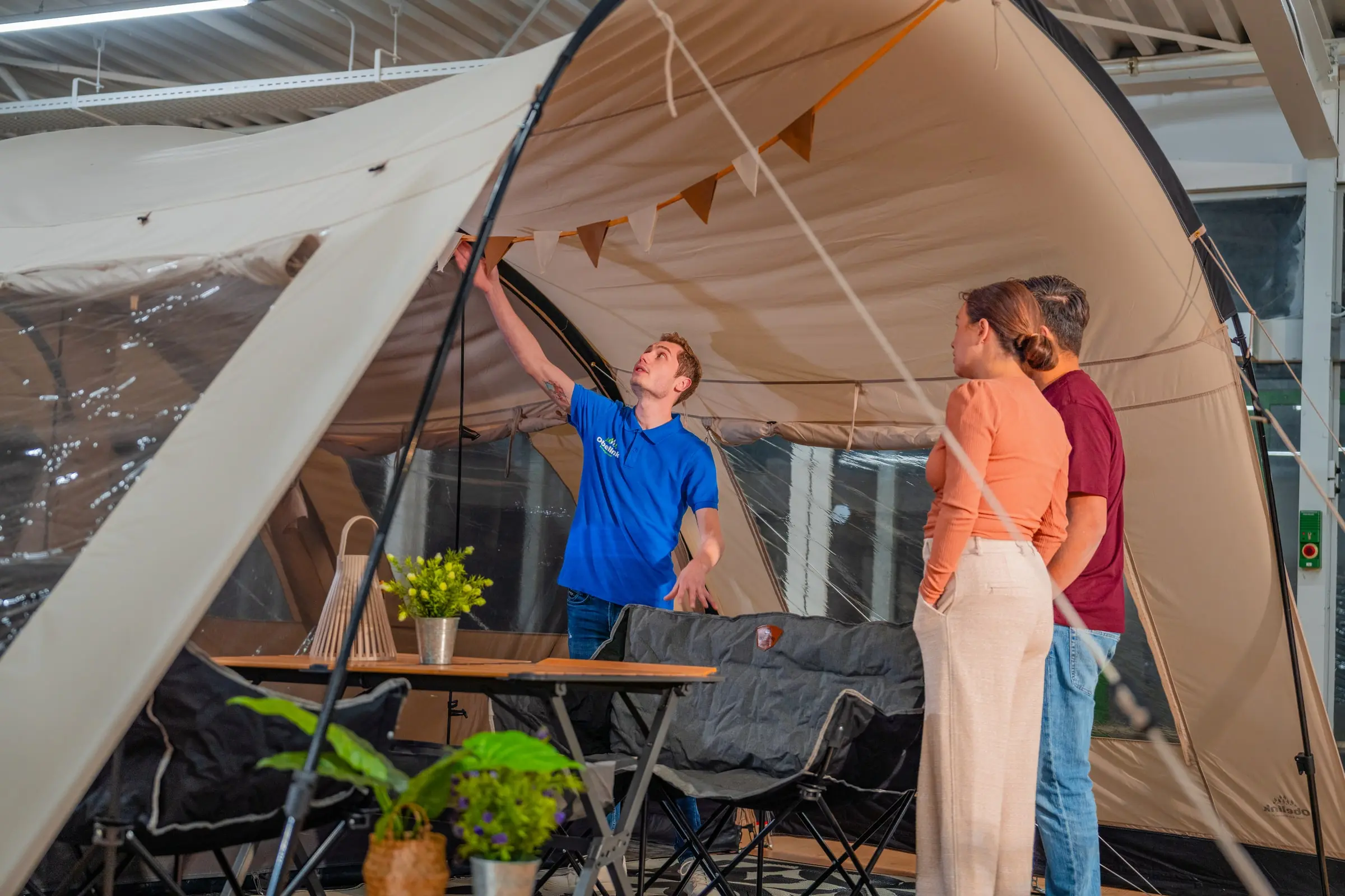 Mitarbeiter in blauem Hemd demonstriert ein großes Familienzelt für Kunden in einem Geschäft, mit Campingtisch und Stühlen innerhalb des aufgebauten Zeltes