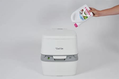 Wasser und Spülflüssigkeit zum Spülwassertank der Campingtoilette hinzufügen Hand hält weiße Flasche mit rosa Deckel über einer weißen tragbaren Toilette vor einem hellgrauen Hintergrund