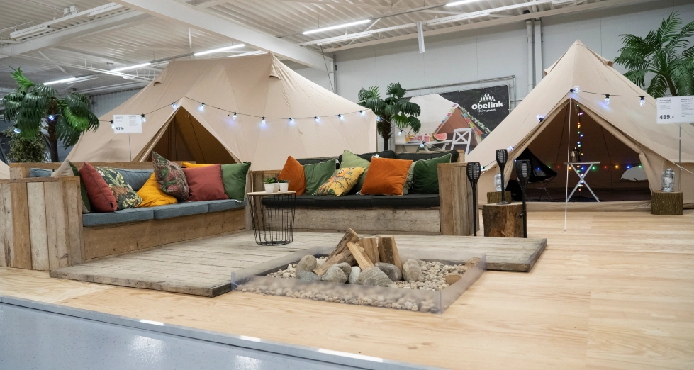 Geschäft mit Sahara-Zelten Luxuriöse beige Glampingzelte mit bunten Kissen auf einer Holzterrasse, dekorative Feuerstelle und festliche Beleuchtung in einer Showroom-Aufstellung