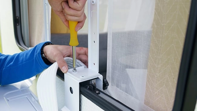 Schritt 4: Wie montiert man eine Split-Klimaanlage Person in blauem Pullover befestigt mit einem gelben Schraubenzieher ein Teil am Fensterrahmen eines Wohnwagens oder Wohnmobils.