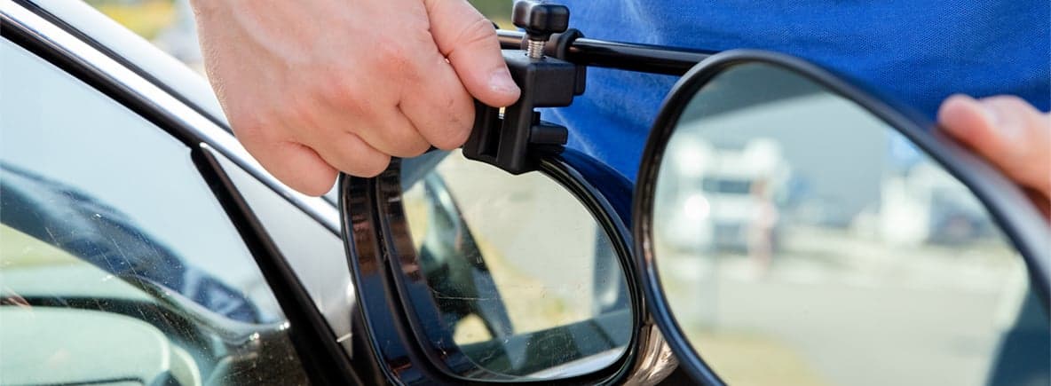 Sicher mit dem Wohnwagen verreisen: Checkliste! Nahaufnahme einer Hand, die einen Wohnwagenspiegel an einem blauen Fahrzeug montiert, mit verschwommenem Hintergrund.