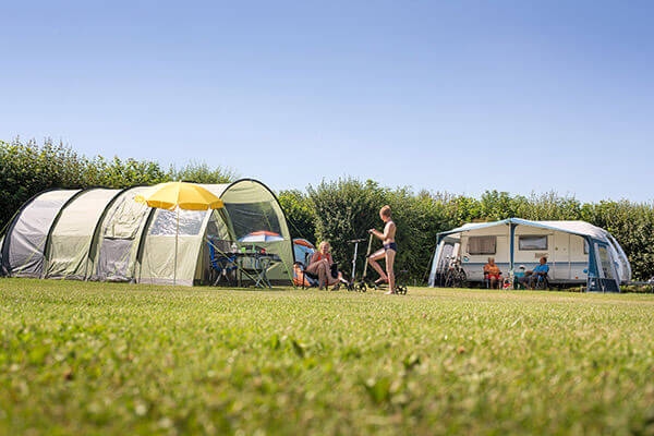Zeltplatz Ferienparkt t Akkertien Geräumiger Campingplatz mit Tunnelzelt und Wohnwagen auf einer grünen Wiese unter blauem Himmel, Menschen entspannen sich an Campingtischen an einem sonnigen Tag.