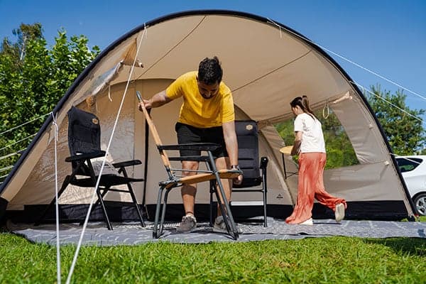 Mann klappt Stuhl vor dem Zelt aus Geräumiges Kuppelzelt mit Vorzelt auf einer Grasfläche, zwei Personen stellen Campingstühle und Tisch unter sonnig blauem Himmel bereit