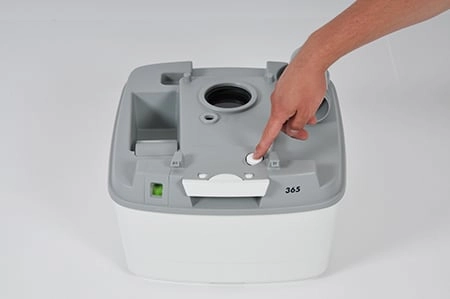 Entlüftungsknopf Porta Potti Tragbare Toilette mit grauem Oberteil und weißem Unterteil, wobei eine Hand den Entlüftungsknopf drückt.